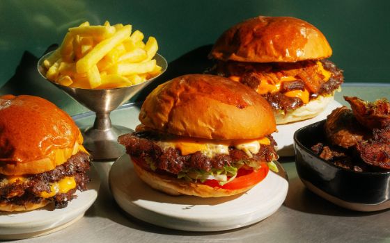 Drei Cheeseburger und Beilagen, angerichtet auf verschiedenen Tellern auf einer Ablage aus Edelstahl, professionell beleuchtet. 