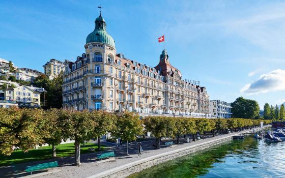 Ein grosses historisches Gebäude mit zwei kleinen Türmen und diversen Verzierungen liegt direkt an einem Fluss mit einer kleinen Baumallee. 
