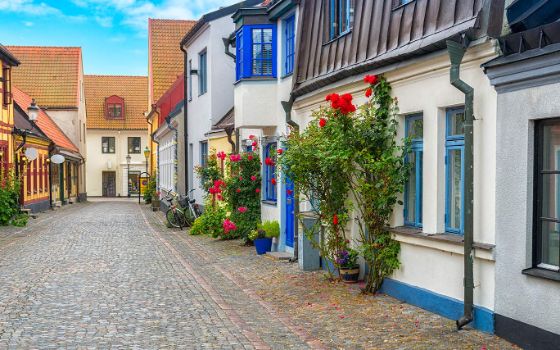 Kleine Häuser mit bunten Fensterläden und Blumen vor den Türen stehen in einer schmalen Kopfsteinpflasterstrasse. 