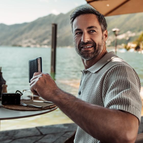 Ein stilvoll gekleideter Mann an einem Esstisch vor einem See hält ein Kartenetui mit einer silbernen Kreditkarte von American Express hoch.