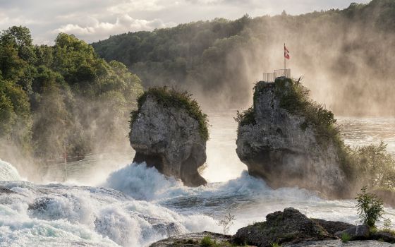 Rheinfall bei Schaffhausen mit Felsen in der Gischt und Schweizer Fahne auf Aussichtsplattform.