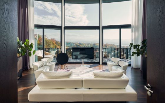 Blick auf eine moderne Sitzgruppe aus hellem Leder und einen Fernseher, dahinter grosse Panoramafenster und ein Balkon mit Aussicht auf Baumwipfel.