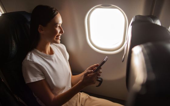Eine lächelnde Frau sitzt mit Smartphone in den Händen auf einem Fensterplatz in einem Flugzeug.