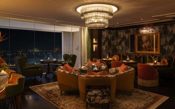 In einem Raum mit bodentiefer Fensterfront stehen Sessel um gedeckte Tische, an der Decke hängt ein grosser Kronleuchter und die Wände haben gemusterte Tapeten mit Bildern. 