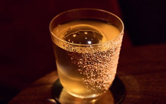 Nahaufnahme eines hellgelben Getränkes mit einem runden Eiswürfel in einem schlichten, runden Glas auf einem Holztisch.