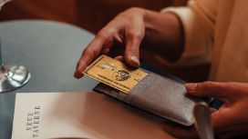 Eine Person hält an einem Tisch im Restaurant ein braunes Lederetui mit einer goldenen Kreditkarte von American Express.