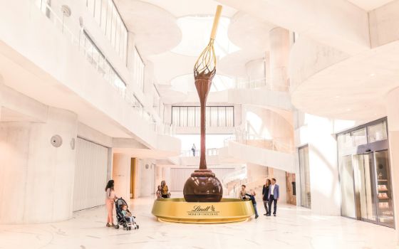 In einem hellen Raum stehen Menschen um einen grossen Schokoladenbrunnen, an der Decke hängt ein übergrosser Rührlöffel, aus dem die Schokolade in einen goldenen Brunnen fliesst. 