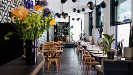In einem Restaurant mit dunklem Fussboden stehen an der Wand und vor den Fenstern Tische und Stühle aus Holz, im Vordergrund steht eine Vase mit Blumen. 