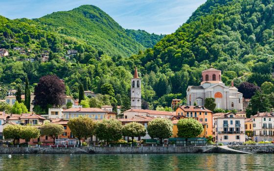 Italienische Kleinstadt mit Kirche und Uferpromenade an einem See vor bewaldeter Berglandschaft. 