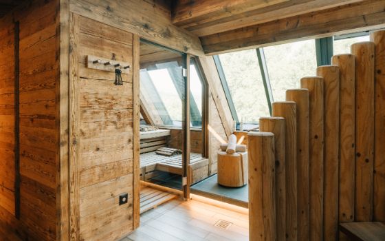 Ein mit Holz verkleideter Raum mit einer kleinen Sauna mit Glastüre in einer Dachschräge mit grossen Fenstern, daneben eine Holztreppe, die nach oben führt.