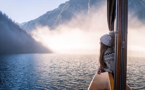 Profil einer Frau im Norwegerpullover mit Wollmütze, die aus dem Fenster eines Sschiffes in eine nebelverhangene Berglandschaft auf einem See schaut. 