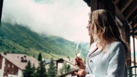 Frau auf einem Balkon mit Glas in der Hand, im Hintergrund Häuser, Berge und Wolken