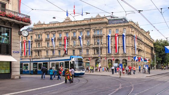 Historisches Bankgebäude mit Schweizer Flaggen an einem belebten Platz, auf dem eine Tram fährt.
