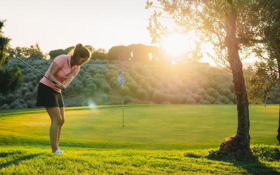 Frau im Rock beim Abschlag auf einem Golfplatz mit mediterraner Bepflanzung im warmen Sonnenlicht. 