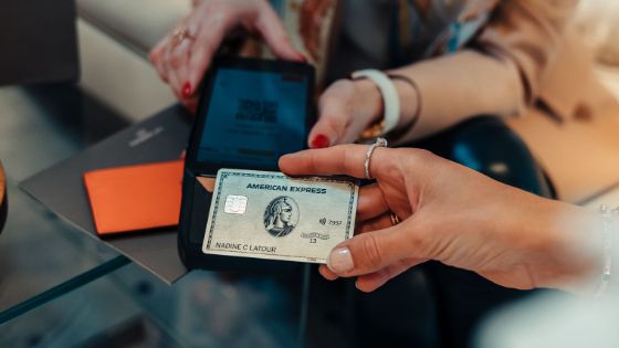 Kontaktlose Zahlung mit einer silbernen Kreditkarte von American Express aus Metall an einem Kartenlesegerät in den Händen einer Verkäuferin in einer Boutique