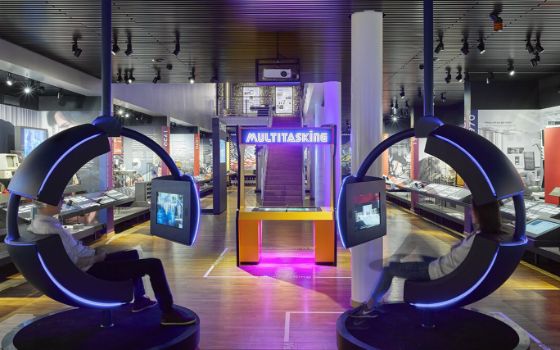 Zwei kreisrunde, beleuchtet Sessel mit integrierten Monitoren stehen in einem grossen Raum, der mit neon-farbigen Lichtern beleuchtet ist.