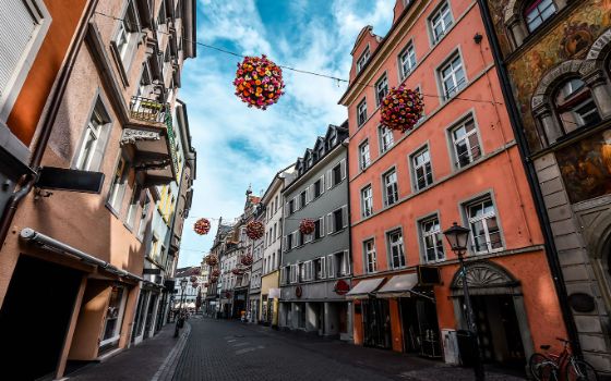 Blick in eine schmale Altstadtstrasse in Konstanz mit farbigen Blumenkugeln, die über der Strasse hängen, und historischen Gebäuden links und rechts.