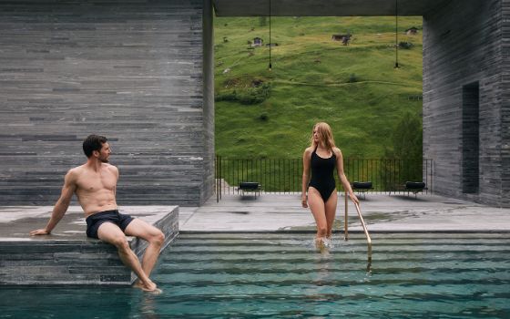 Eine Frau in schwarzem Badeanzug steig in einen grossen Außenpool, während ein Mann in Badehose am Rand sitzt. 