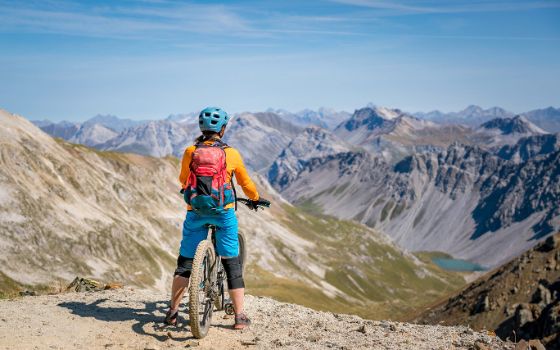Eine Frau mit Mountainbike steht an einem Bergvorsprung und schaut in die Landschaft.